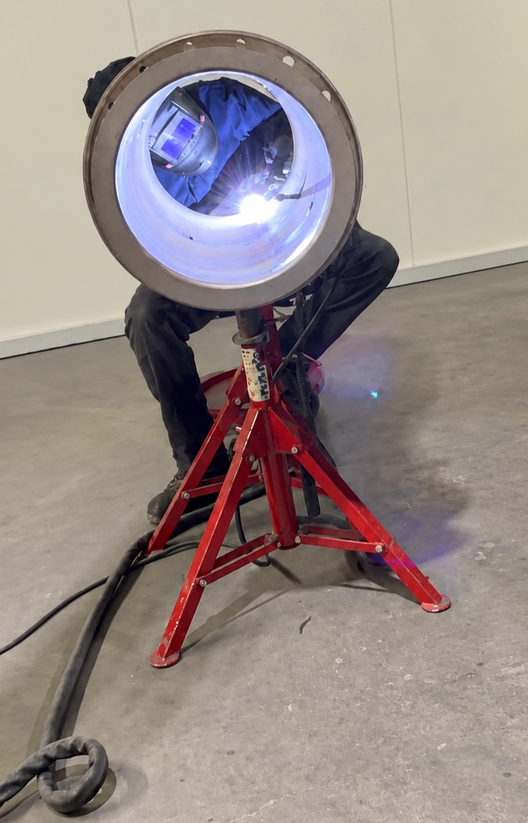 A welder works on a cylindrical part resting on a red stand in a workshop.